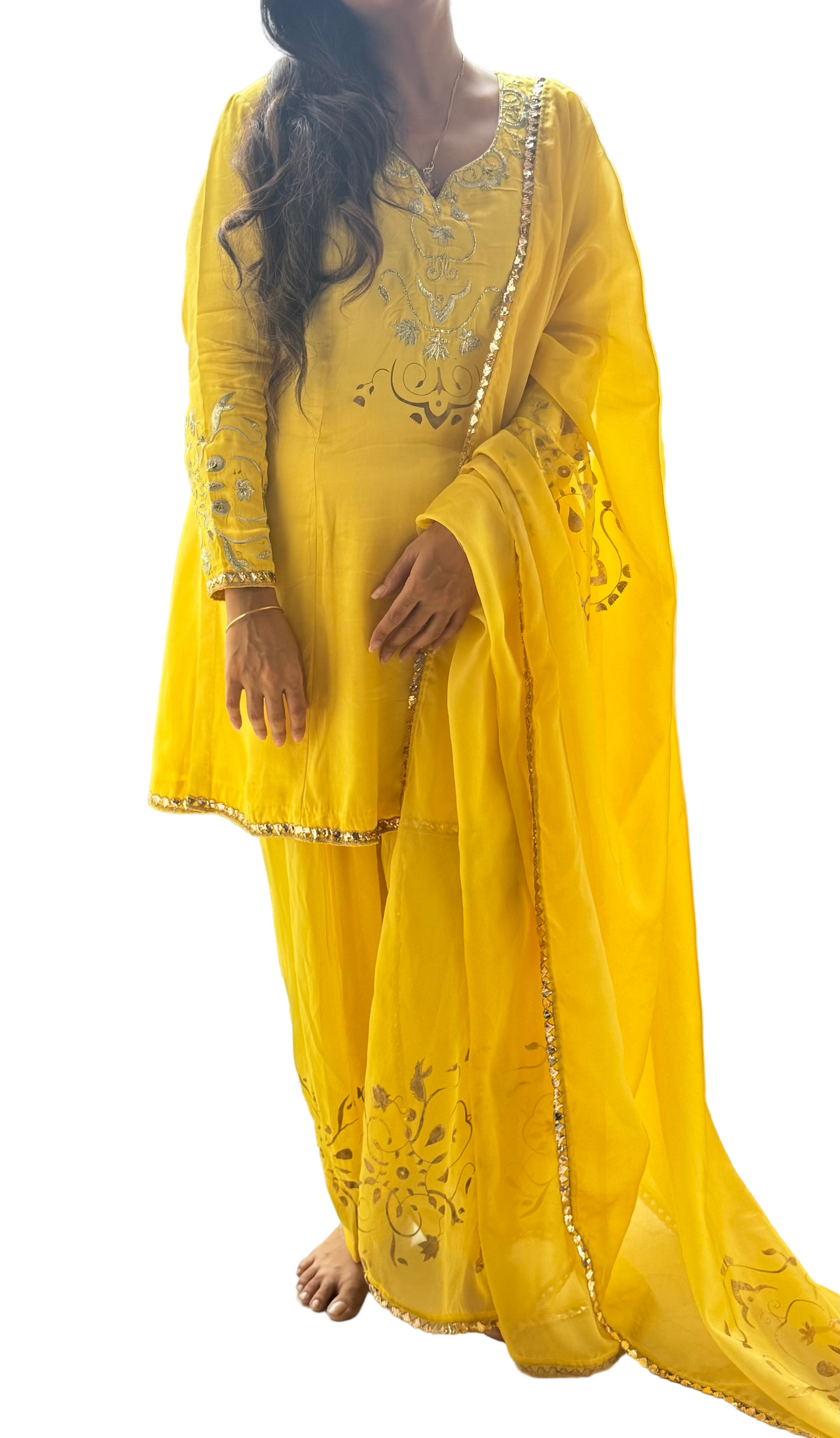 Person wearing a yellow traditional outfit with intricate designs, standing by a window with greenery outside.