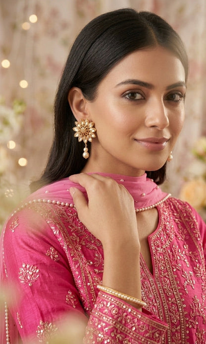 Woman wearing a pink traditional outfit with gold embroidery and jewelry against a blurred floral background.