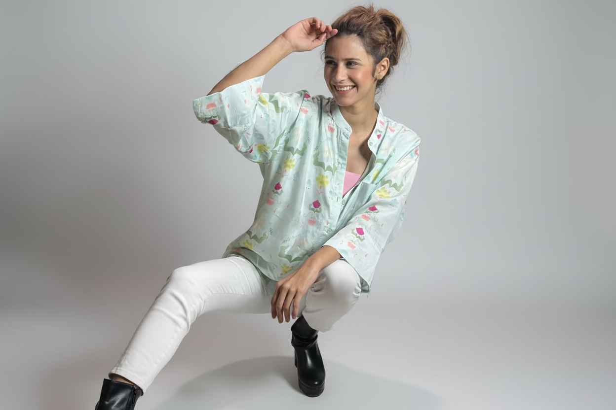 Woman sitting outdoors wearing a floral blouse and white pants, surrounded by plants.