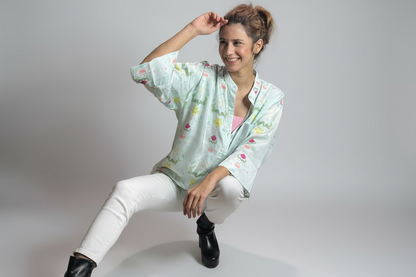 Woman sitting outdoors wearing a floral blouse and white pants, surrounded by plants.